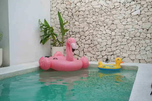 Inflatable pink flamingo and yellow duck lifebuoys float on the water in a pool in summer. Stone background with houseplants decoration on the side. Holiday and vacation concept.