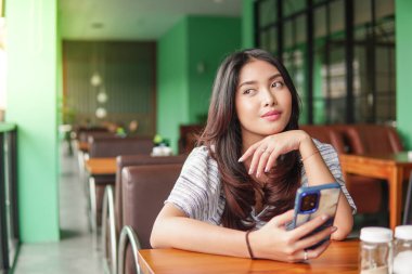 Hayal kuran genç Asyalı bir kadın kahvaltı için bir restoranda oturmuş, elinde akıllı bir telefonla düşüncelere dalmış.