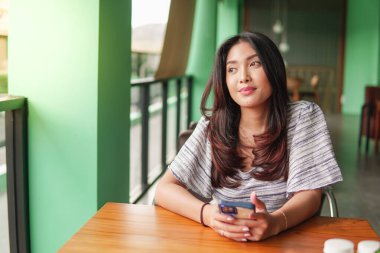 Gülümseyen Asyalı bir kadın elbise giyiyor ve mutlu hissediyor. Kahvaltıda elinde akıllı bir telefonla restoranda oturuyor.