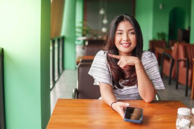 Genç ve güzel Asyalı bir kadın kahvaltı yapmak için restoranda otururken sağ tarafını işaret eden akıllı telefon kullanıyor.