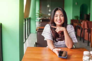 Genç ve güzel Asyalı bir kadın kahvaltı yapmak için restoranda otururken sağ tarafını işaret eden akıllı telefon kullanıyor.