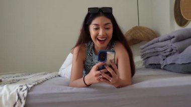 Heyecanlı genç Asyalı kadın, sosyal medyayı kontrol etmek için akıllı telefon aygıtı kullanırken yatağa uzanıyor, internetten satın alıyor, internetten ürünler sipariş ediyor. Tatil yaşam tarzı kavramı