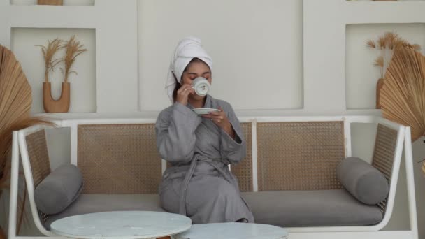 アジアの若い女性は朝のコーヒーや紅茶をソファーに座ってリラックスして灰色のバスローブを着ています ホリデーレジャーコンセプト — ストック動画