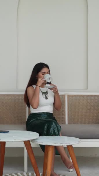迷人的亚洲女人一边坐在沙发上 一边举杯喝着早茶或咖啡饮料 一边享受假期和放松的感觉 — 图库视频影像