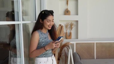 Gülümseyen genç Asyalı kadın sosyal medyayı kontrol etmek için akıllı telefon kullanıyor, internetten ürünler alıyor ve sipariş veriyor. Tatil yaşam tarzı kavramı