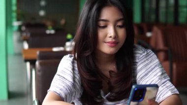 Gülümseyen Asyalı kadın bir restoranda oturmuş kahvaltıyı beklerken mesaj atıyor, sosyal medyada ve internette geziniyor.
