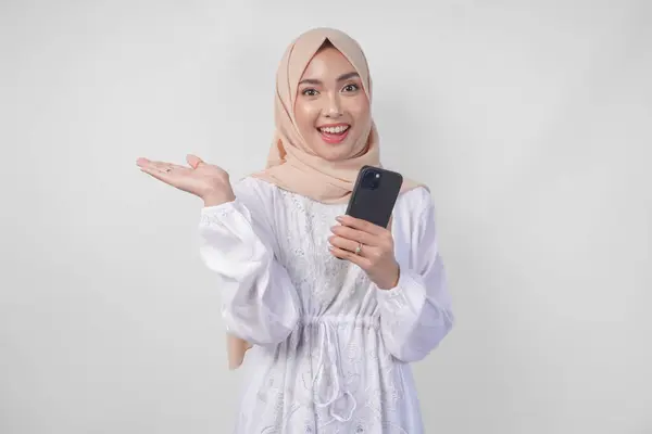 stock image Smiling Asian woman wearing hijab and white dress using smartphone while pointing to the copy space on her right side, standing over isolated white background. Ramadan and Eid Mubarak concept