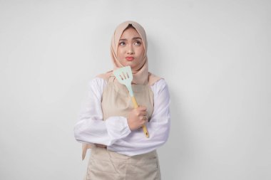 Tesettür ve krem rengi önlüklü düşünceli genç Asyalı Müslüman kadın elinde spatula ve mutfak aletleri, hangi yemek menüsünü pişireceğine kafa yoruyor.