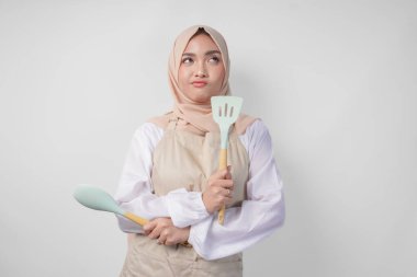 Tesettür ve krem rengi önlüklü düşünceli genç Asyalı Müslüman kadın elinde spatula ve mutfak aletleri, hangi yemek menüsünü pişireceğine kafa yoruyor.