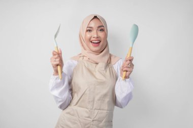Spatula ve mutfak aletleri tutarken kameraya gülümseyen peçe ve krem önlüklü heyecanlı Asyalı genç Müslüman kadın.