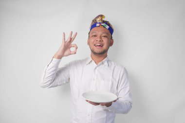 Beyaz gömlekli ve geleneksel başlıklı mutlu genç Balili adam elinde fotokopi yeri olan boş bir tabak tutarken yemeğin ne kadar güzel olduğunu ifade etmek için nefis bir el hareketi yapıyor.