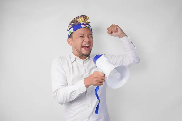 stock image Energetic Balinese man in white shirt and traditional headdress shouting at megaphone while raising clenched fist, isolated by white background