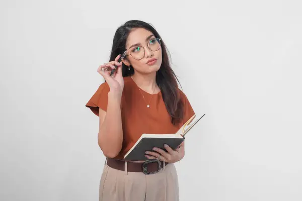 Thoughtful Young Asian Woman Wearing Brown Shirt Eyeglasses Holding Book stockbilde