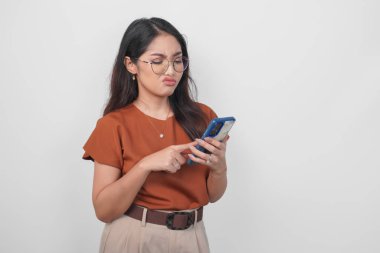 Kahverengi gömlekli ve gözlüklü genç Asyalı kadın, beyaz arka planla izole edilmiş akıllı telefonu elinde tutan sarkık dudaklarından rahatsız oluyor..