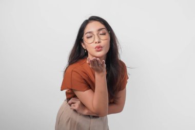 Kahverengi gömlekli ve gözlüklü tatlı Asyalı kadın beyaz arka planda dudakları somurtkan dudaklarıyla öpücük yolluyor..