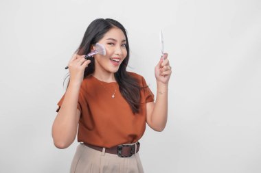 Ayna ve makyaj fırçası tutan güzel genç bir kadın yeni kozmetik ürünler kullanarak allık sürmek için izole edilmiş beyaz arka plan hazırlıyor..