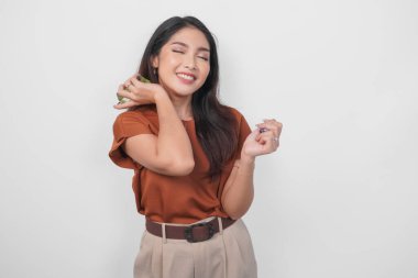 Kahverengi tişörtlü güzel bir Asyalı kadın elinde parfüm şişesi ve boynuna sprey sıkarak beyaz arka planla izole edilmiş..