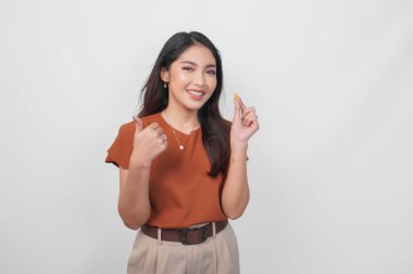 Kahverengi gömlekli neşeli Asyalı kadın elinde ilaç hapı tutarken beyaz arka planla izole edilmiş baş parmak hareketlerini gösteriyor..