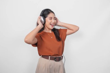 Kahverengi gömlekli neşeli genç Asyalı kadın kablosuz kulaklık kullanarak müzik dinliyor ve eğleniyor..