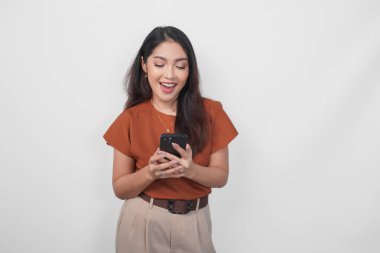 Kahverengi gömlek giyen çekici genç Asyalı kadın elinde akıllı telefonunu tutarken gülümsüyor..