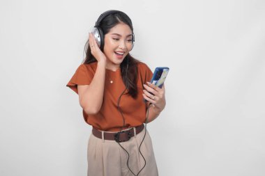 Kahverengi tişörtlü neşeli genç Asyalı kadın kablosuz kulaklık kullanarak akıllı telefondan müzik dinliyor..