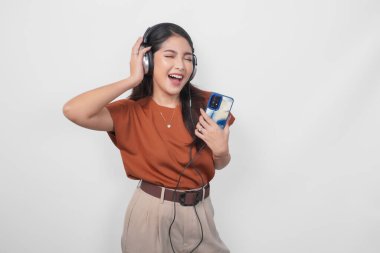 Kahverengi tişörtlü mutlu bir Asyalı kadın kablosuz kulaklık kullanarak gülümsüyor ve akıllı telefondan müzik dinliyor..