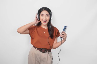 Kahverengi tişörtlü mutlu bir Asyalı kadın kablosuz kulaklık kullanarak gülümsüyor ve akıllı telefondan müzik dinliyor..