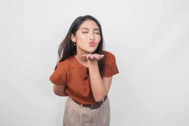 Kahverengi gömlekli tatlı Asyalı kadın beyaz arka planda izole edilmiş kameraya dudaklarını büzerek öpücük yolluyor..