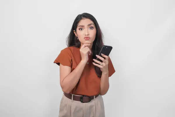 白い背景の上に隔離されたスマートフォンを保持しながら 脇に見える茶色のシャツを着た思慮深い若い女性 ロイヤリティフリーのストック画像