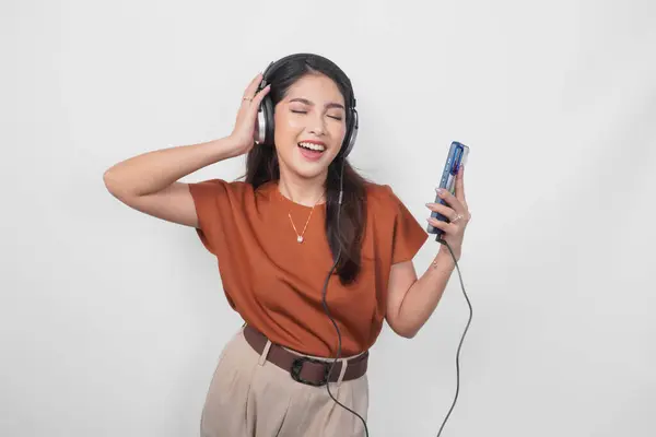 ブラウンのシャツで幸せな若いアジアの女性 笑顔とワイヤレスヘッドフォンを使用してスマートフォンから音楽を聞く ストックフォト
