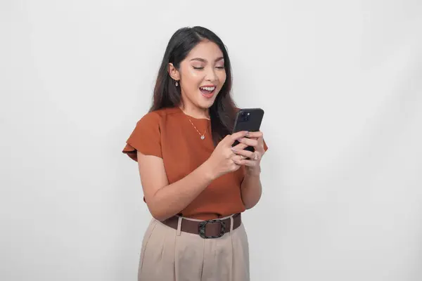 白い背景で隔離された彼女のスマートフォンを使用しながら 茶色のシャツで楽しいアジアの女性 ストック画像