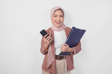 Genç Asyalı Müslüman kadın tesettüre sarılmış, elinde belge kitabı ve akıllı telefon ile kameraya gülümsüyor..