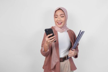 Tesettürlü güzel Asyalı Müslüman kadın, beyaz arkaplanla izole edilmiş internete mesaj atarken veya internette gezinirken bir belge tutuyor..