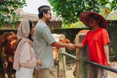 Genç Asyalı Müslüman çift sığırcıyla tokalaşıyor ve kurbanlık sığır almak için para veriyorlar. Kurban Bayramı kavramı.