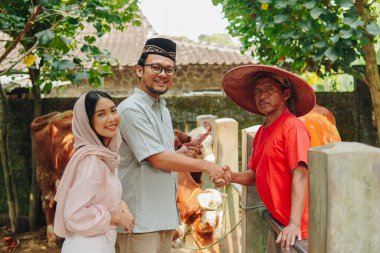 Genç Asyalı Müslüman çift sığırcıyla tokalaşıyor ve kurbanlık sığır almak için para veriyorlar. Kurban Bayramı kavramı.