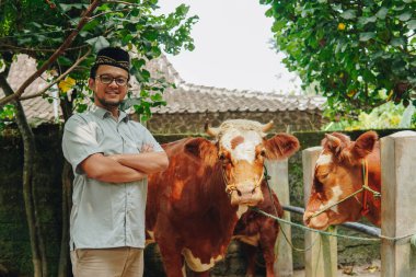 Kurban etmek için çiftlik hayvanlarının önünde gülümseyen, kendinden emin, Asyalı Müslüman bir adam. Kurban Bayramı kavramı.