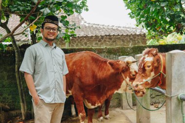 Kurban etmek için çiftlik hayvanlarının önünde gülümseyen, kendinden emin, Asyalı Müslüman bir adam. Kurban Bayramı kavramı.