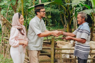 Mutlu Asyalı Müslüman çift, sığırcıyla tokalaşıyor ve kurbanlık hayvan keçileri almak için para veriyor. Kurban Bayramı kavramı.