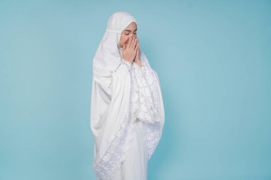 Asyalı ciddi bir müslüman kadın dua elbisesi giyip elleri ile dua ediyor, mavi arka planla izole edilmiş. Hac kavramı.