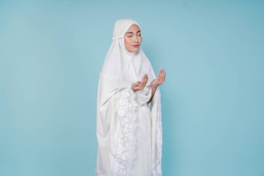 Asyalı ciddi bir müslüman kadın dua elbisesi giyip elleri ile dua ediyor, mavi arka planla izole edilmiş. Hac kavramı.