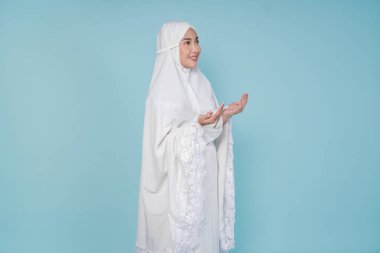 Asyalı ciddi bir müslüman kadın dua elbisesi giyip elleri ile dua ediyor, mavi arka planla izole edilmiş. Hac kavramı.