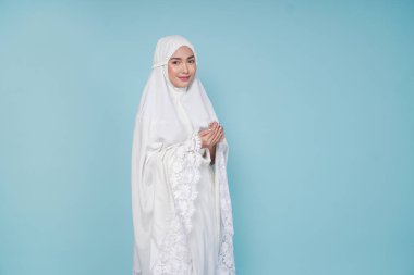 Asyalı ciddi bir müslüman kadın dua elbisesi giyip elleri ile dua ediyor, mavi arka planla izole edilmiş. Hac kavramı.