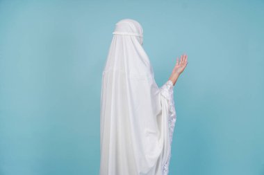 Asyalı müslüman bir kadının dua elbisesi giymiş, mavi arka planla izole edilmiş, elleri dua eden pozu. Hac kavramı.
