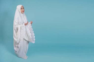 Gülümseyen Asyalı Müslüman kadın dua elbisesi giyip elleri ile dua ediyor, mavi arka planla izole edilmiş. Hac kavramı.