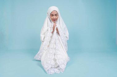 İbadet cüppeli genç Asyalı Müslüman kadın mavi arka planla izole edilmiş gözleri kapalı dua ediyor. Hac kavramı.