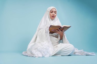 İbadet cüppeli, ciddi bir Asyalı genç Müslüman kadın izole edilmiş mavi arka planda oturup Kur 'an okuyor. Hac kavramı.