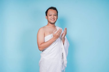 Hacı giymiş gülümseyen Asyalı Müslüman adam mavi arka planla izole edilmiş el hareketleriyle dua ediyor. Hac kavramı.