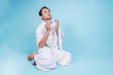 İhramda oturan ve el hareketleriyle dua eden ciddi Asyalı genç Müslüman bir adam mavi arka planla izole edilmiş. Hac kavramı.