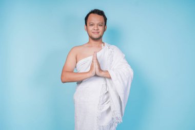 İhram giyen çekici genç Asyalı Müslüman adam mavi arka planla izole edilmiş geleneksel dini selamlaşmaları işaret ediyor. Hac kavramı.