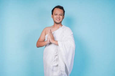 İhram giyen çekici genç Asyalı Müslüman adam mavi arka planla izole edilmiş geleneksel dini selamlaşmaları işaret ediyor. Hac kavramı.